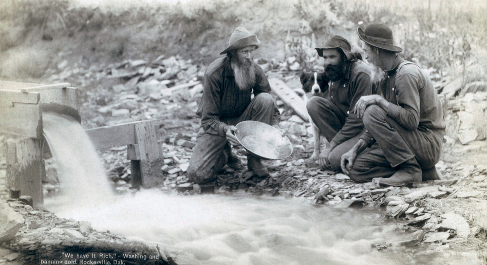 Three men, with dog, panning for gold in a stream in the Black Hills of South Dakota in 1889