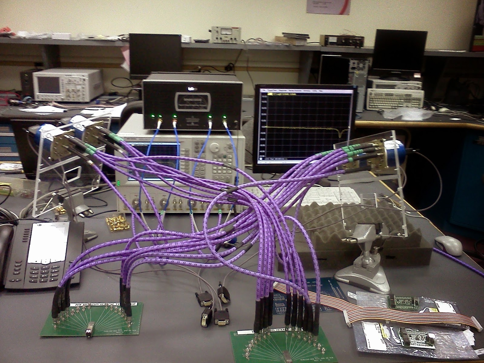 Various lab devices in the background with partially loaded SP12T Radiall switches mounted to a plexiglass frame in the foreground.
